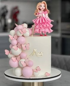 a white and pink cake with a doll on top, surrounded by small balloons in the shape of butterflies