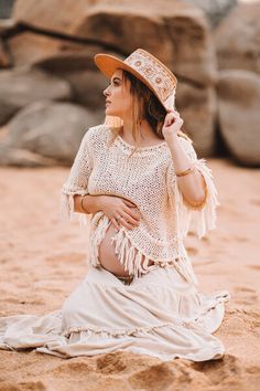 a woman sitting in the sand wearing a hat and holding her hand on her hip