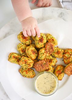 a baby reaching for some food on top of a white plate with a dipping sauce