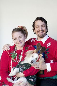 a man and woman in ugly christmas sweaters holding a small dog wearing reindeer antlers