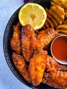 a plate with chicken wings, french fries and dipping sauce on it next to lemon wedges