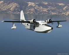 an airplane flying over the water with mountains in the background