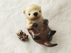 two ceramic otters sitting on top of a white sheet next to each other and one is holding its baby