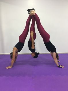 two women doing handstands on purple mats in a yoga studio with one holding the other upside down