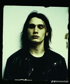 a man with long hair wearing a leather jacket and looking at the camera while standing in front of a white wall