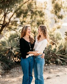 two women standing next to each other in front of some cactus bushes and trees with their arms around each other