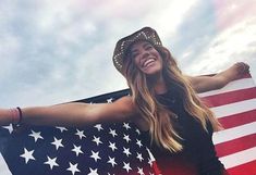 a woman holding an american flag in front of a cloudy sky with her arms outstretched