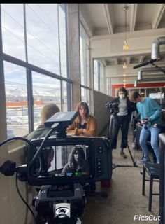 a camera set up in front of a window with people standing and sitting around it