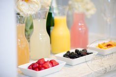 various drinks are lined up on a counter with flowers in vases and fruit bowls