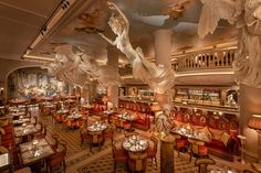 the interior of a fancy restaurant with red chairs and gold tables, angel sculptures on the ceiling