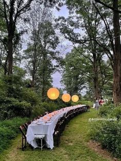 a long table set up in the middle of a forest with lanterns hanging above it