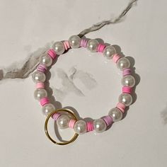 two bracelets with pink and white beads are on a marble countertop next to a pair of gold rings