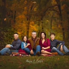 a family sitting on the grass in front of trees with fall foliages behind them