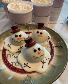 three frosted snowmen on a plate with cups of hot chocolate in the background