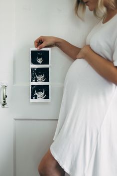 a pregnant woman in white dress standing next to a wall with three pictures on it