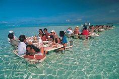 a group of people sitting at a table in the ocean eating food and drinking water