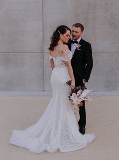 a bride and groom standing next to each other in front of a concrete wall holding bouquets