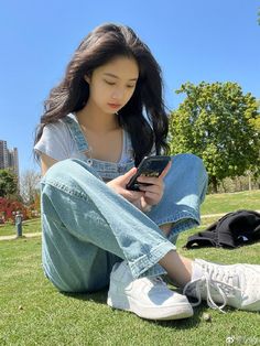 a young woman sitting on the grass looking at her cell phone while she's reading something