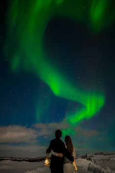 two people are standing in the snow under an auroral sky filled with green lights