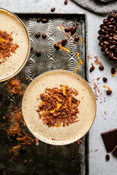 two glasses filled with dessert sitting on top of a metal tray next to coffee beans