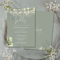 two wedding cards with white flowers and string lights in the background on top of a gray cloth