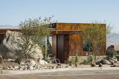 a house made out of rusted metal and surrounded by desert plants, rocks and trees