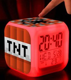 a hand is pointing at the time on a cube shaped alarm clock with an orange and red design