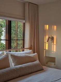 a bed with white sheets and pillows in front of a window that has light coming through the windows