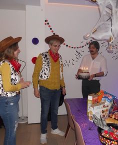 three people standing around a table with a cake on it and one person wearing a cowboy hat