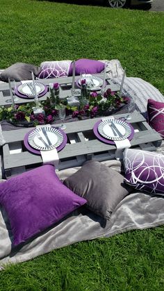 a picnic table set up with purple and white pillows, plates and flowers on it