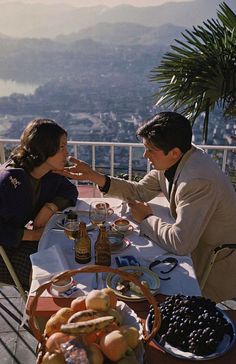 a man and woman sitting at a table with food on it, overlooking the city