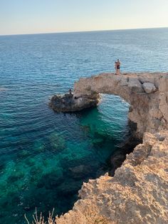 two people are standing on the edge of a cliff over looking the ocean and rocks