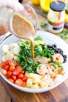 someone pouring dressing over a bowl of food on a cutting board with olives, tomatoes and shrimp