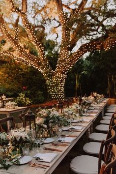 a long table set up with place settings for dinner under a tree filled with lights