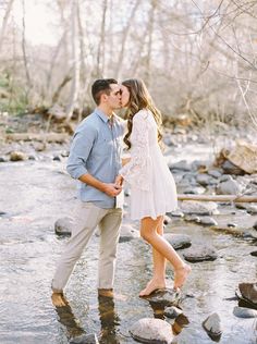 a man and woman kissing in the water on rocks next to a river with trees