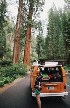 a woman standing in the back of an orange van on a road surrounded by trees