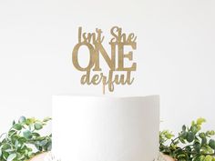 a white cake with gold lettering on top and greenery in the backgroun