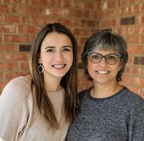 two women standing next to each other in front of a brick wall and smiling at the camera