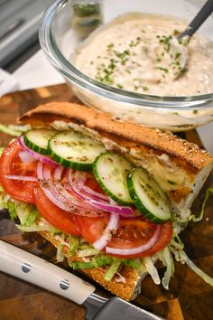 a sandwich with cucumbers, tomatoes and onions on a cutting board next to a bowl of dressing