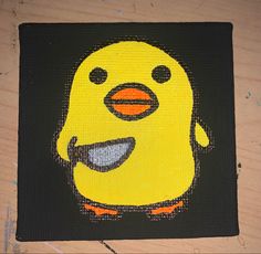 a painting of a yellow bird on a black square with orange feet and mouth, sitting on a wooden surface