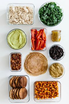 an array of different foods in containers on a white surface, including carrots, lettuce, beans, and hummush