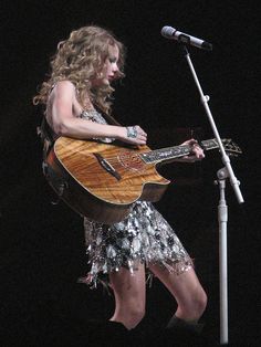 a woman holding a guitar while standing next to a microphone on top of a stage