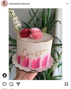 a hand holding a cake with pink frosting and flowers on top, in front of a plant