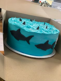 there is a blue cake with black sharks on it in a cardboard box that has been opened