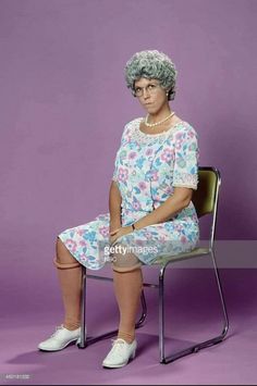 an older woman sitting on a chair with her legs crossed and wearing white shoes, in front of a purple background