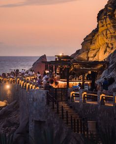 people are sitting at tables next to the water and rocks on the beach as the sun sets