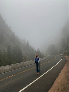 a woman standing on the side of a road in the middle of foggy mountains