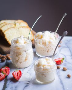 two small jars filled with whipped cream and strawberries on a marble table next to sliced bread