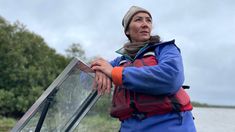 a woman standing next to a body of water wearing a blue jacket and red backpack