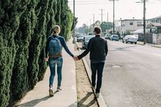 two people holding hands walking down the sidewalk next to a street lined with tall trees